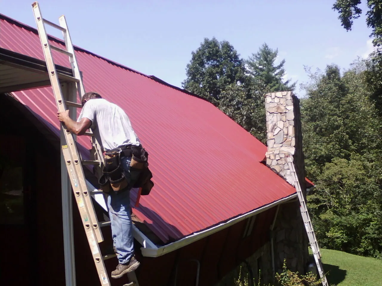 Roofer on a ladder beside a red metal roof and stone chimney — Soffit & Fascia Repair by Roofing Brothers in Shade Gap
